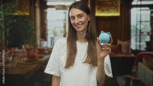 Schilderij op canvas Young woman holding blue piggybank and smiling in livingroom by dining table and sofa; saving money optimism frugality