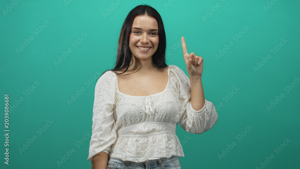 Fototapeta premium Woman smiling and pointing index finger upward in studio, wearing white blouse and jeans against teal backdrop; confidence clarity.