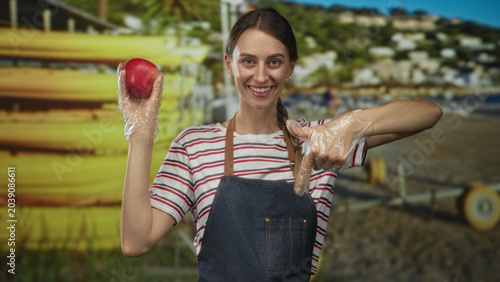 Принти на полотні Woman holds red apple with gloved hand and points finger to apple on beach; healthy eating cheerful