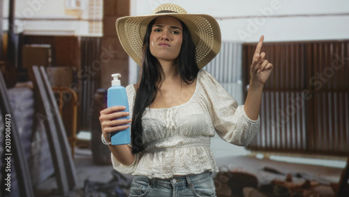 Obraz na płótnie Young woman in straw hat and white blouse holding blue sunscreen bottle points index finger upward in building, displeased expression; sun safety warning