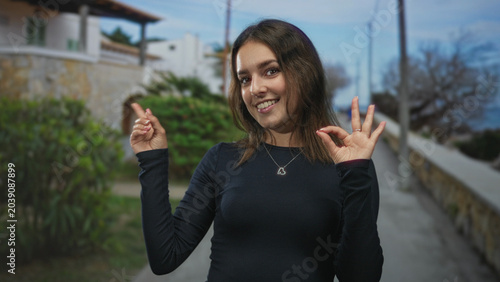 Foto Woman pointing finger and making ok gesture on street, wearing black top and heart necklace, smiling at camera; playful confidence