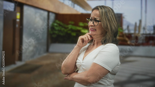 Photos Senior hispanic woman with grey hair in white top and glasses, hand on chin grimacing near a building entrance and outdoor plaza; disgust