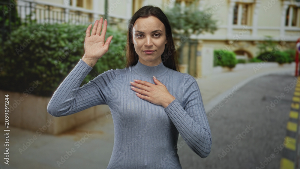 Fototapeta premium Woman standing with hand on chest on street by building, eyes closed and calm facial expression while wearing ribbed sweater; calm reflection.