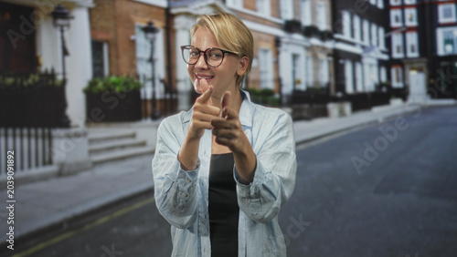 Foto Woman points finger directly at camera on a quiet urban street, smiling and wearing glasses and a casual denim shirt in front of townhouse facades; playful confidence