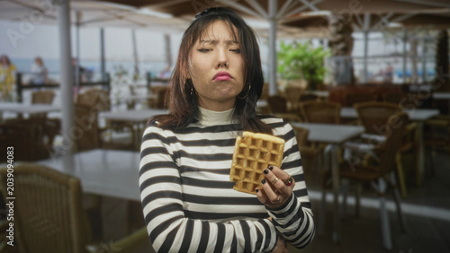 Foto Woman holding waffle, arms crossed and frowning while wearing a striped sweater in a seaside cafe building; annoyance hesitation