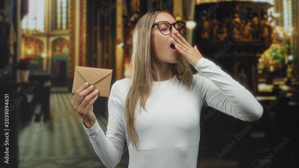 Fototapeta premium Young woman holding a kraft envelope yawning with hand to mouth inside an ornate church building filled with pews and altarpieces; tiredness reflection.