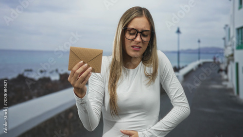 Obraz na plátně Woman wearing glasses holding brown envelope and clutching stomach while grimacing on a seaside street beside a pier walkway; discomfort bad news