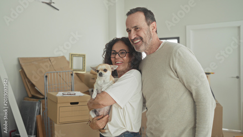 Εκτύπωση καμβά Man and woman embrace while woman cradles small dog and man waves hand amid stacked moving boxes and picture frame in building; moving day joy