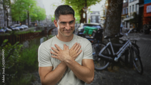 Obraz na płótnie Man with bare hands crossed over chest, smiling with palms pressed to sternum on a canal street beside a parked bicycle and riverside walkway; gratitude warmth connection