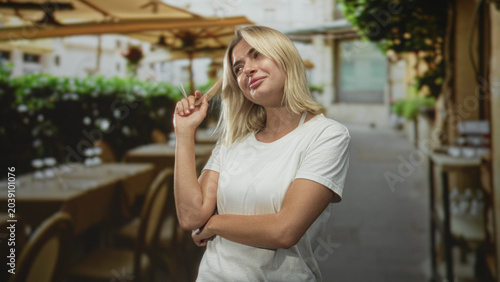 Obraz na płótnie Woman twirls blonde hair with fingers in street cafe, arms crossed and smirking; playful confidence
