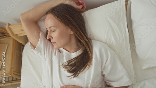 Принти на полотні Woman lying in bed with arm above head and hand on stomach on white sheets beside a rattan nightstand in building; serenity rest