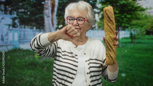 Tableau sur toile Elderly woman holds a long baguette and displays thumb down gesture beside a building in a park setting; disapproval nostalgia