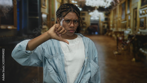 Photography Woman with glasses and braided hair showing thumbs down gesture in museum building gallery, denim shirt visible and raised hand; disapproval critique
