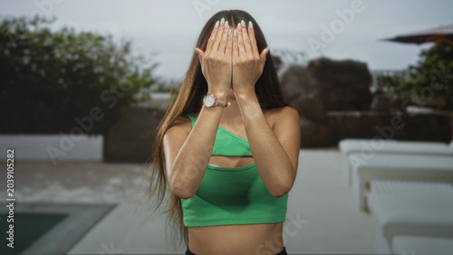фотографія Woman with hands covering eyes at outdoor pool wearing green crop top and watch, midriff visible; embarrassment hiding