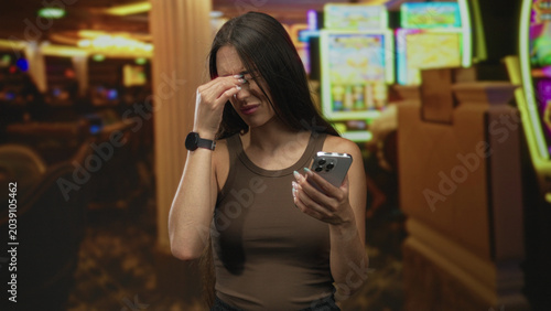 Canvas-taulu Woman pinches bridge of nose and rubs eyes while holding smartphone in casino building with slot machines in view; frustration loss