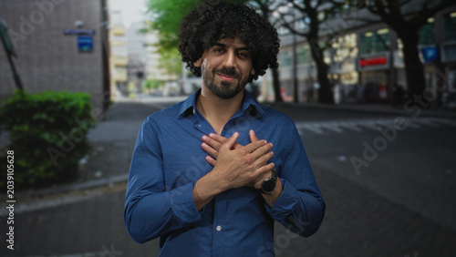 Valokuva Man with hands crossed over chest on a city street wearing a blue shirt, curly hair and beard visible, smiling gently; gratitude sincerity calm