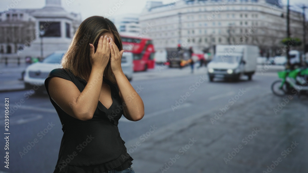 Fototapeta premium Woman covers face with hands on a busy city street with passing bus and van amid midday traffic and blurred buildings; vulnerability anxiety.