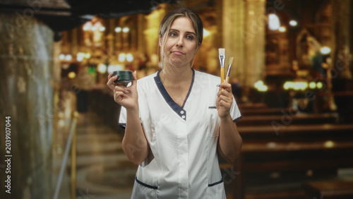 Foto Woman in white coat holds jar and paintbrushes up with eyes closed and lips pursed standing in building aisle; resignation doubt