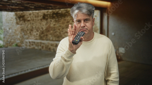 Foto Man holding tv remote control with right hand, wearing cream knit sweater and slight frown while standing by stone accent wall and sliding glass door in studio; quiet boredom