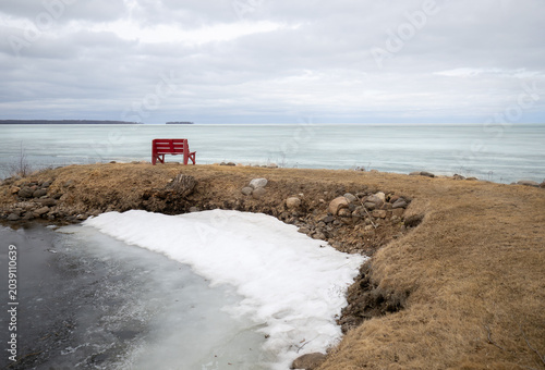 A red bench sits out on a peninsula, as the ice of the lake and overcast clouds add a cold feeling to the day