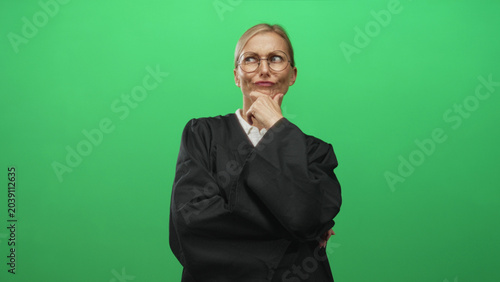Картината върху платно Woman judge in black robe with hand on chin and glasses in a studio green screen setting, thinking gesture; thoughtful reflection