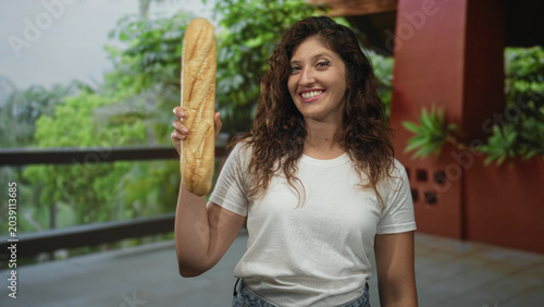 Obraz na plátně Young hispanic woman smiles and holds a baguette aloft in right hand on hotel balcony inside building; joy friendly warmth