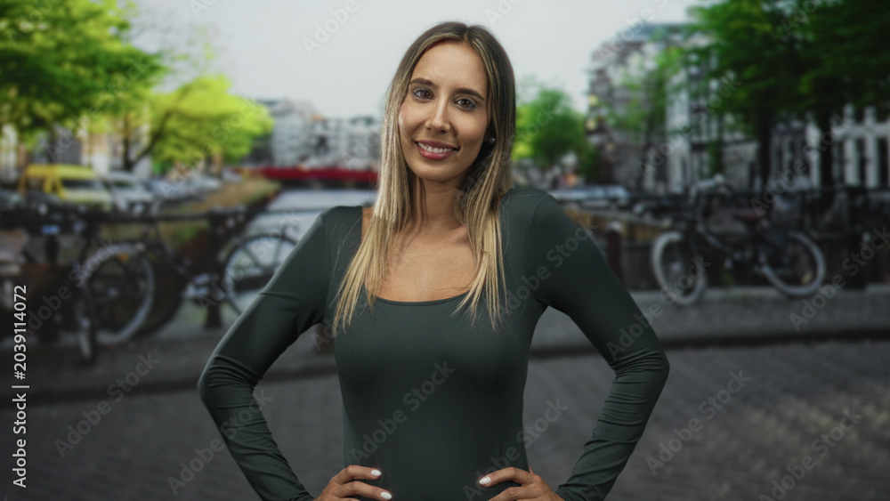 Fototapeta premium Woman with hands on hips showing shoulders at street canal in amsterdam; confidence travel exploration.
