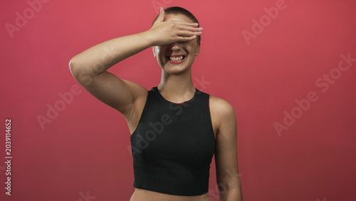 Tablou pe pânză Woman with short hair wearing black crop top holds her right hand covering eyes while smiling, showing bare midriff against a red studio backdrop; playful shyness