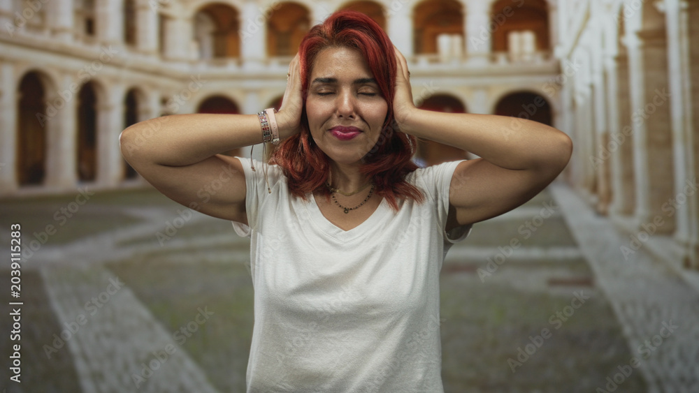 Fototapeta premium Woman with red hair covering ears with fingers in an old town stone building courtyard, eyes closed and smiling; playful defiance.