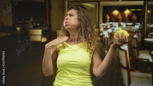 Fényképezés Woman in yellow top holding yellow piggybank, touching throat with hand and gesturing while looking upward in restaurant; savings worry annoyance