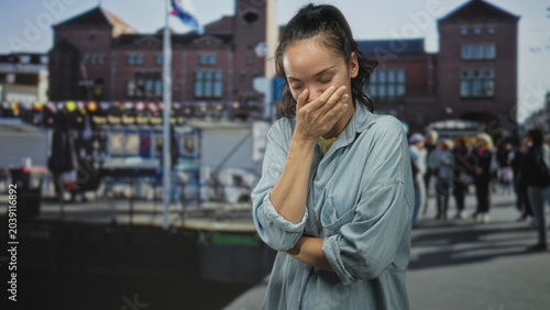 Bild auf Leinwand Young woman covering mouth with hand on a busy street market in front of brick building; amusement introspection