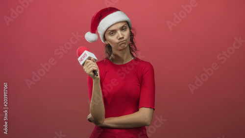 Canvas Print Young woman wearing santa hat and red shirt holding microphone with arms crossed and pouting in studio; skepticism holiday irony