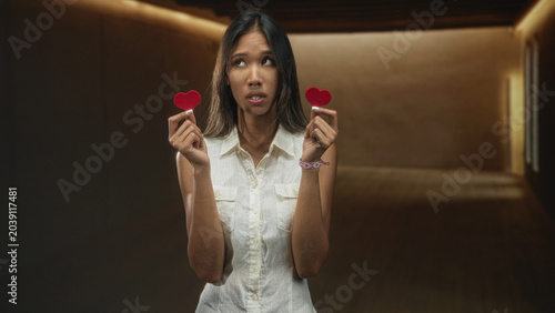 Tableau sur toile Woman holding two red heart props between hands, comparing hearts while standing in a dim building corridor; uncertainty longing