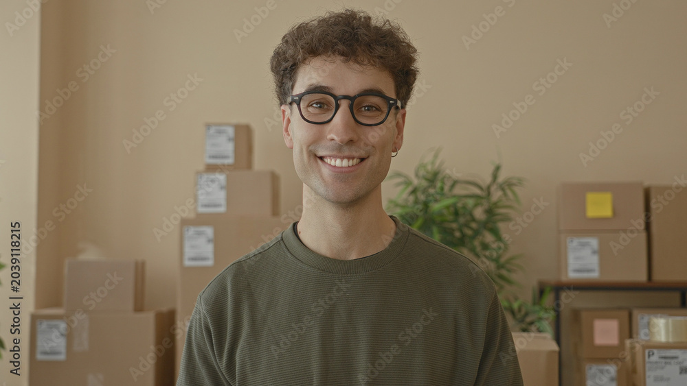 Fototapeta premium Young man smiles at camera amid stacked shipping boxes and parcels on shelves in a building; friendly service.