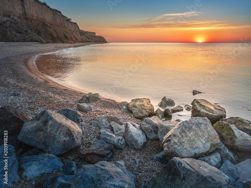 Curved sea coast with cliff and stones in water and rising sun