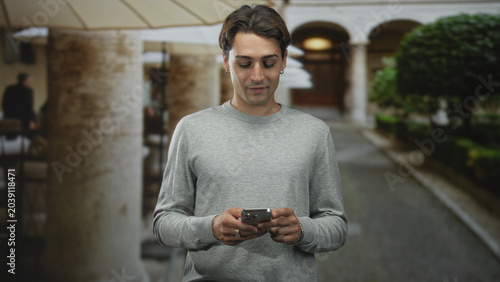 Foto Man holding smartphone and glancing sideways in a building colonnade courtyard with umbrella seating and clipped topiary; thoughtful quiet moment