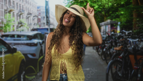 Obraz na plátne Woman tipping straw hat with index finger, yellow crochet top and midriff exposed on a street lined with bicycles and parked cars; summer fun playful