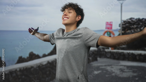 Foto Man teenager with outstretched bare arms, smiling with eyes closed, breathing sea air on a street promenade by rocky coast; joy freedom optimism