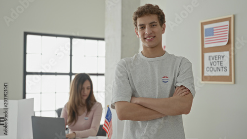 Φωτογραφία Man with arms crossed wearing i voted sticker beside seated woman at laptop and american flag in a polling building; civic duty pride