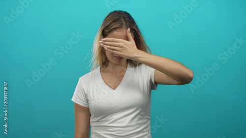 Foto Woman covering her eyes with hand in studio against turquoise background, wearing white v neck t shirt, frontal pose; shy privacy