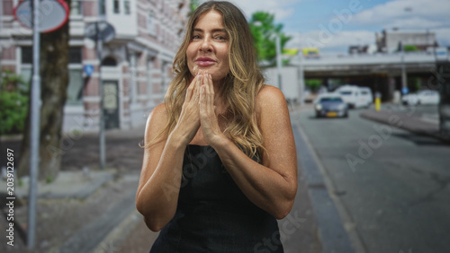 Foto Woman middle aged with hands pressed together at chin in pleading prayer gesture, standing on street near sidewalk, overpass and parked cars; gratitude