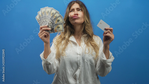 Canvas Print Middle age woman holding fan of cash and a white credit card in hand, pouting lips and slightly tilting head against a blue studio backdrop; playful confidence