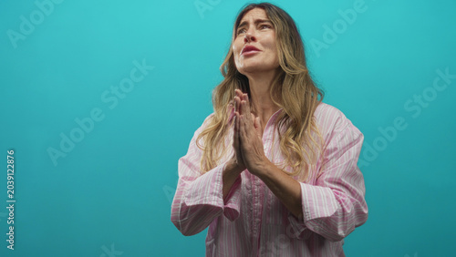 Quadro em tela Woman with hands clasped in prayer looking upward against turquoise studio backdrop; hopeful earnest plea