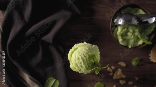 This flat lay video captures a single vibrant scoop of green pistachio ice cream garnished with a fresh mint leaf, set against a dark rustic wooden background with a waffle cone nearby.