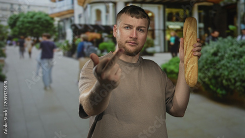 Photography Young caucasian man with beard holding a long baguette and showing middle finger directly to camera on a busy urban street lined with shops; anger defiance provocation