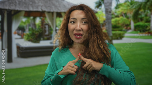 Slika na platnu Woman touching index fingers together at a resort cabana near pool wearing green sweater and red nails; hesitant reflection decision