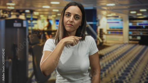Billede på lærred Woman points finger at camera and touches neck in busy airport check in area wearing white t shirt; assertive warning