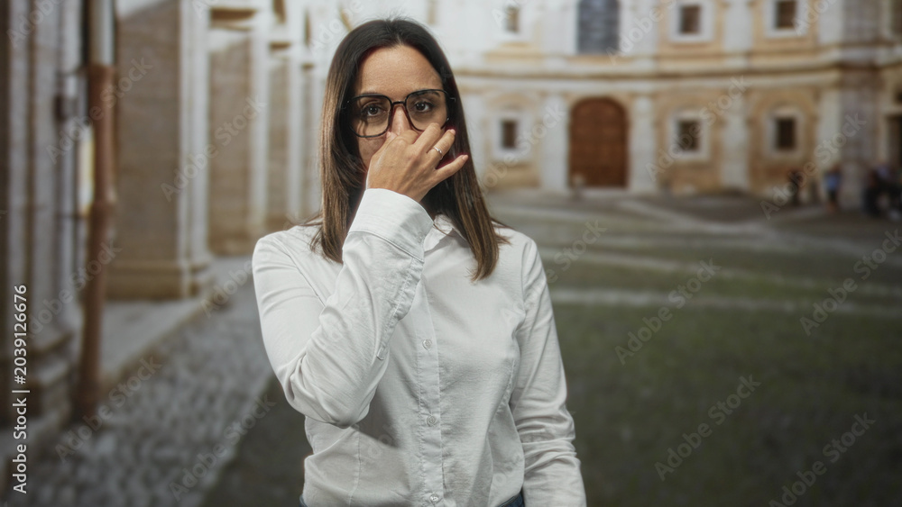 Fototapeta premium Woman pinches nose with hand and raises other palm near face in historic building courtyard; disgust avoidance.