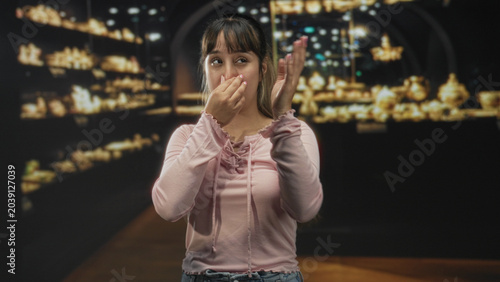 фотографія Woman pinches nose and grimaces, holding raised hand near face among glass display cases and gold artifacts in a museum; disgust