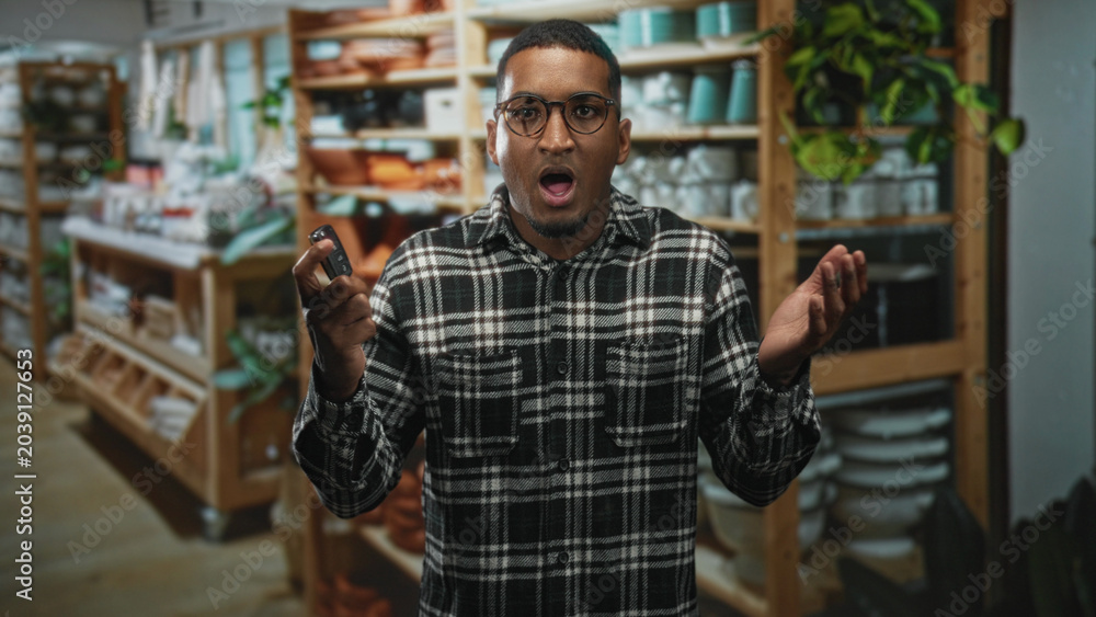 Obraz premium Man holding car keys with mouth open and hands raised in a shrug gesture standing in an aisle of a home goods shop building; confusion.
